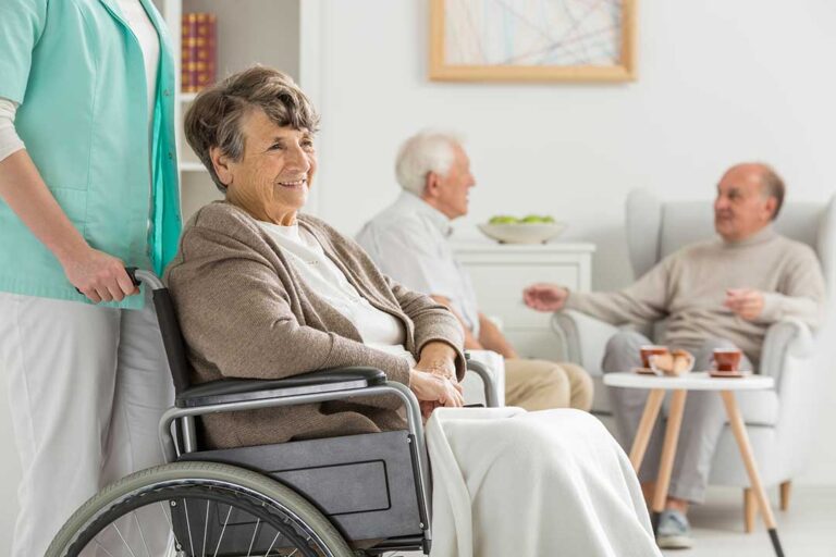 An older adult painting at a table in a sunlit common room with other residents nearby.